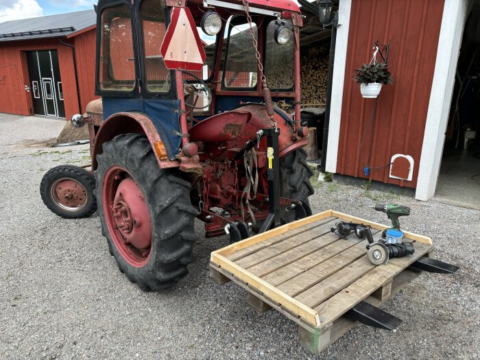 En röd traktor med gafflar på baklyften och en pall med redskap, inklusive en borrmaskin och en vinkelslip, framför en röd byggnad.