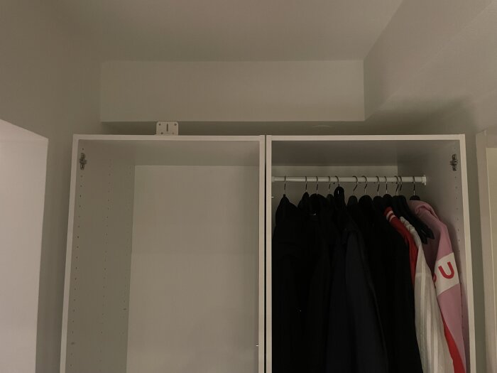 Två vita garderober, den vänstra är tom medan den högra innehåller kläder på galgar. Ett vinkeljärn ovanpå garderobernas mitt vid taket syns.