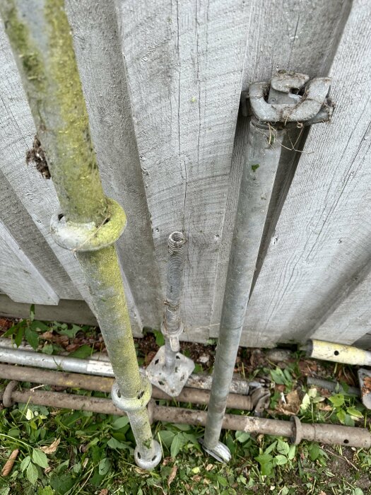 Metallrör av olika längder och former med skruvfästen och kopplingsmekanismer lutade mot ett träplank, omgivna av gräs och löv.