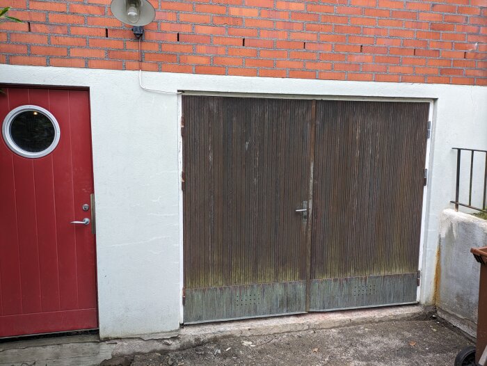 Garageport från 1960-talet i mörkt trä bredvid en röd dörr med runt fönster, byggnad med vit puts och röd tegelfasad ovanför.