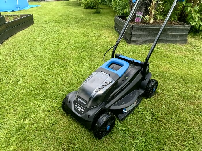 Batteridriven gräsklippare av märket Scheppach 20v med blå och svarta detaljer står på en nyklippt gräsmatta med odlingslådor i bakgrunden.