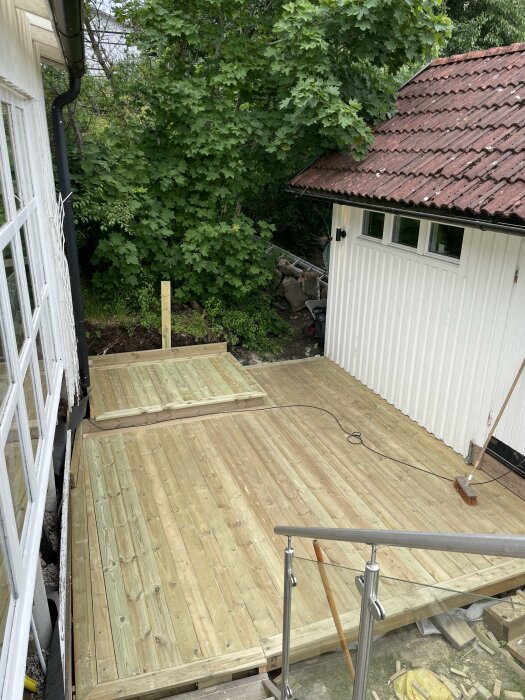 Träveranda under konstruktion bredvid ett vitt hus och en träbod med rött tak. Trappsteg och räcken finns på plats. Området omges av grönska.