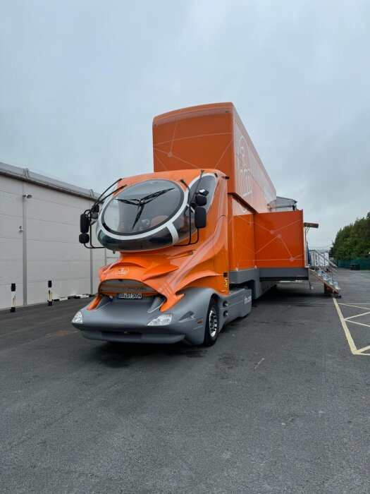 Stor orange futuristisk lastbil parkerad på ett industrifält med ramper utfällda på sidan.