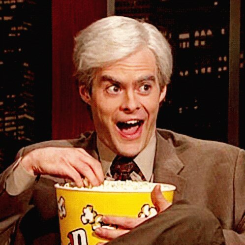 En man i kostym med grått hår äter popcorn och ser förvånad eller glad ut.
