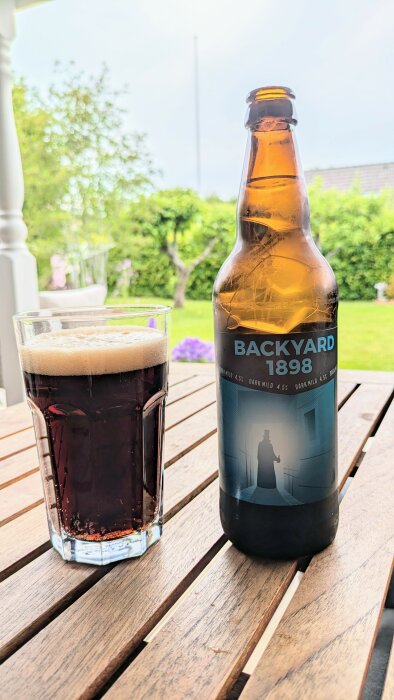 Ölflaska med etikett "Backyard 1898" och ett glas upphällt mörkt öl på ett träbord utomhus, med en grön trädgård i bakgrunden.