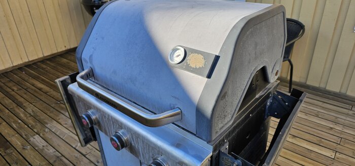 Äldre Landmanngrill med rost och smuts på locket, termometer monterad på sidan, placerad på en träterrass framför en träpanelvägg.