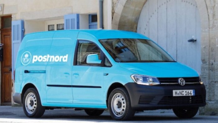 En blå PostNord-fyrhjulsdriven skåpbil parkerad framför en byggnad med ljusblå fönster.