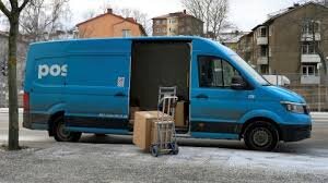 Blå leveransbil med öppen sidodörr, två kartonger och en lastkärra utanför, parkerad på en gata med bostadshus i bakgrunden.