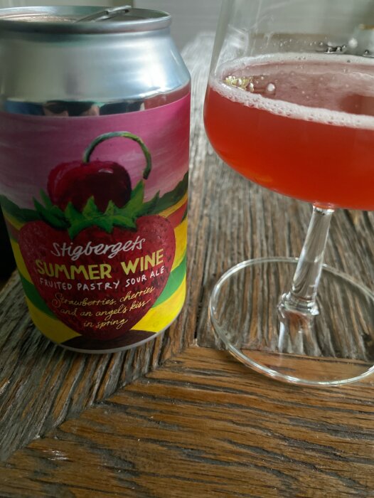 En burk Stigbergets Summer Wine sour ale bredvid ett halvtomt glas med röd öl på ett träbord.