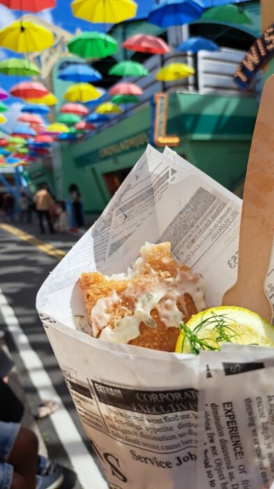 Fish and chips with lemon served in a paper cone, held up against a street adorned with colorful umbrellas hanging above.