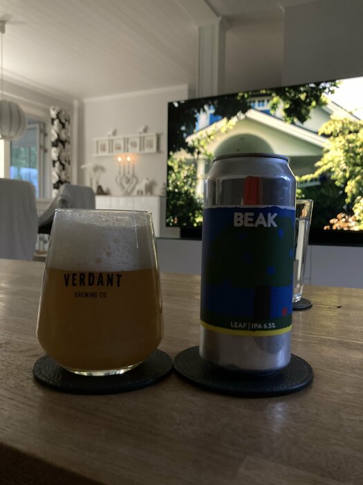 En öl från Beak Brewery står på ett bord bredvid ett ölglas från Verdant Brewing Co. En tv-skärm och en inredning syns i bakgrunden.