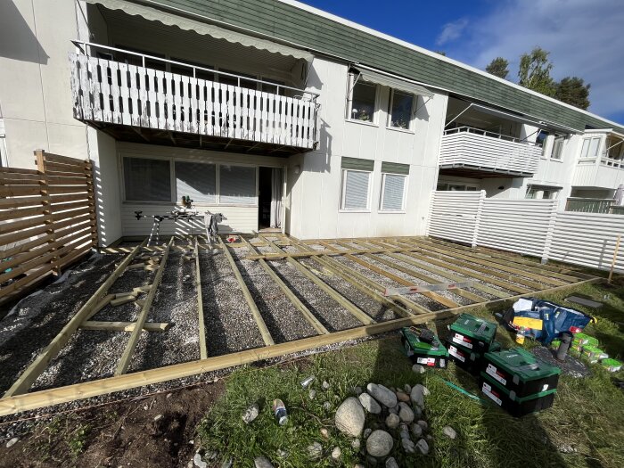 Pågående bygge av en trädäckstruktur i en trädgård, med träreglar placerade på marken framför ett vitt tvåvåningshus med balkong. Verktyg och byggmaterial syns bredvid.