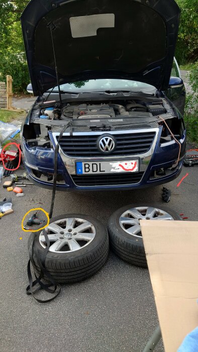 Bil under reparation med huven öppen, verktyg och två bildäck på marken. En borrmaskin och en domkraft syns också i bilden.