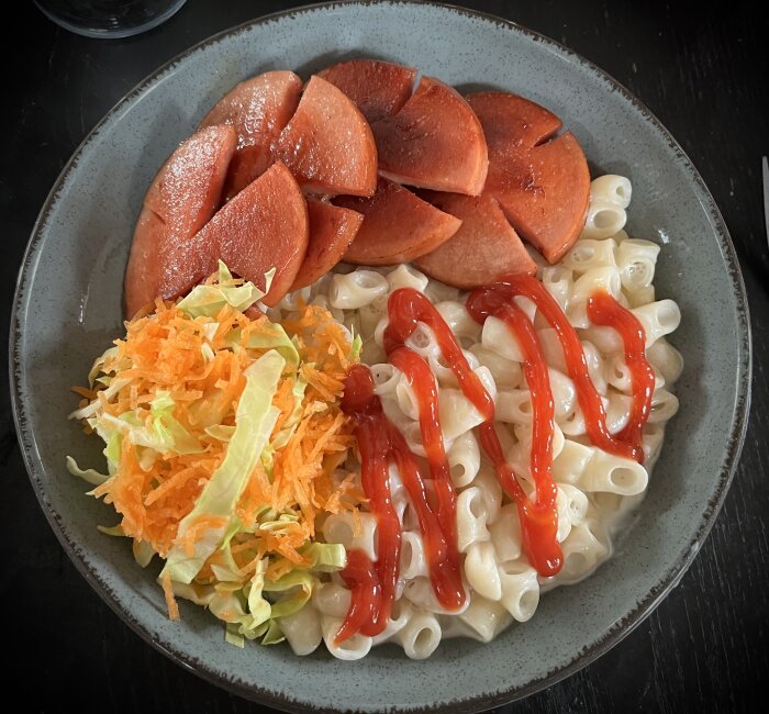 En tallrik med stuvade makaroner toppade med ketchup, stekt falukorv och rårivna grönsaker (morot och vitkål).