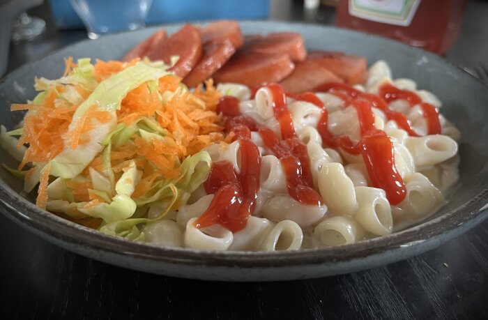 En tallrik med stuvade makaroner täckta med ketchup, skivad korv och en sallad med strimlad morot och vitkål.