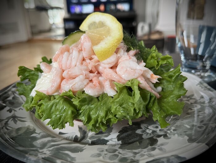 Räkmacka med salladsblad, räkor, majonnäs, citronskiva och dill på ett dekorativt fat, placerad på ett bord.