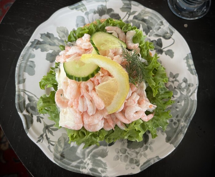 Räkmacka med sallad, citronskiva, gurka och dill på en mönstrad tallrik.