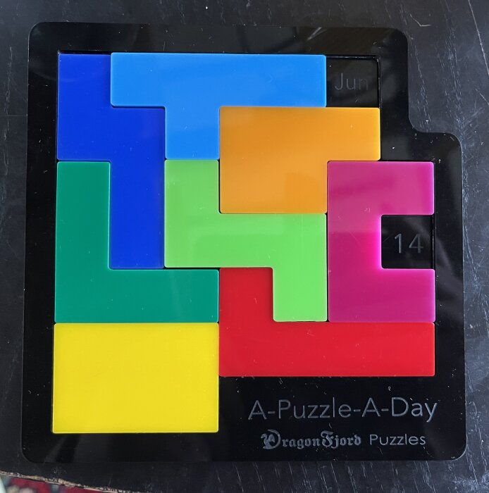 Ett pussel med färgglada bitar i olika former och färger monterade på en svart ram märkt "A-Puzzle-A-Day" och "DragonFjord Puzzles". Dagens datum är 14 juni.