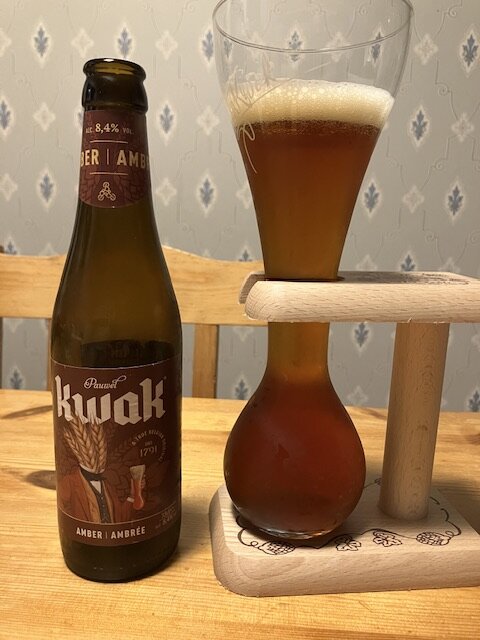 En flaska öl av märket Kwak och ett fyllt ölglas med träställning på ett träbord.