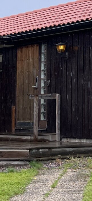 Träytterdörr med glasparti på sidan vid huvudingång till villa, mörkbrun träfasad med utomhuslampa ovanför dörren.
