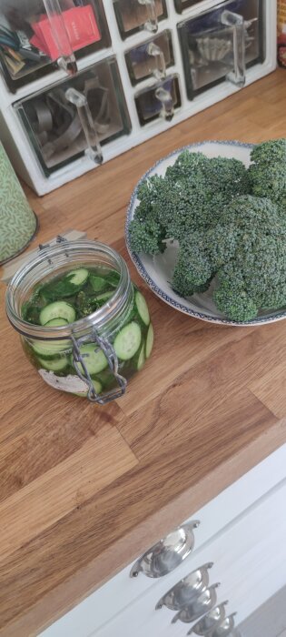 En glasburk med snabbsaltad skivad gurka och en tallrik med färsk broccoli på en köksbänk i trä.