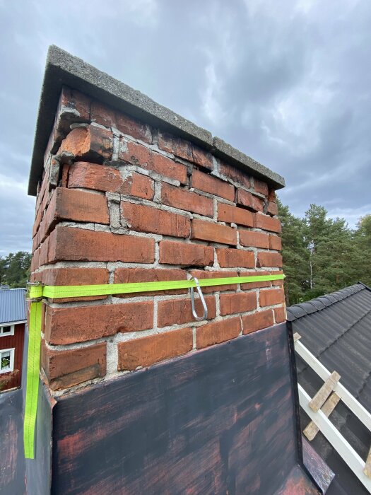 En äldre tegelmurad skorsten med slitage på tegelstenarna, omgiven av mörka takplåtar, med grönt band fäst för att stabilisera strukturen.