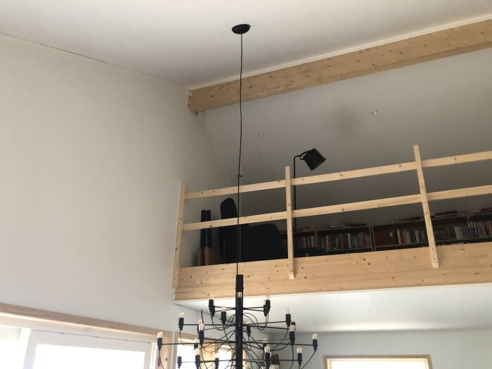 Loft med träbalkong, svart fåtölj, vägglampa och bokhylla. Svart takkrona i förgrunden.