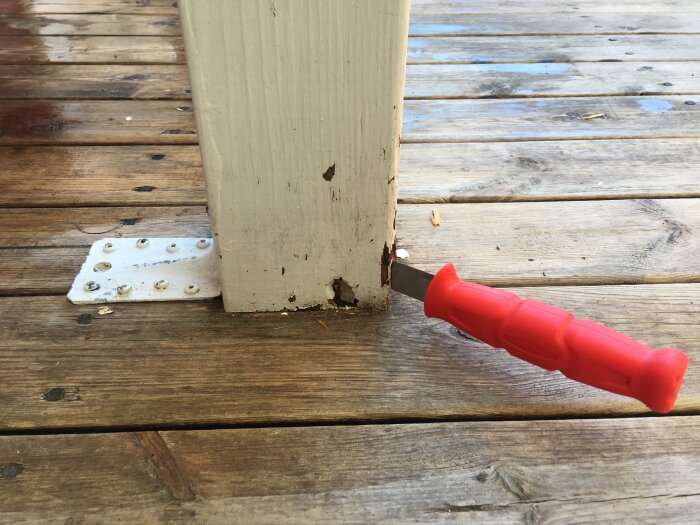 Nedre delen av en vitmålad stolpe med rötskador, fastskruvad i trädäck. En kniv med ett rött handtag är instucken i den flagnande och ruttna ytan på stolpen.