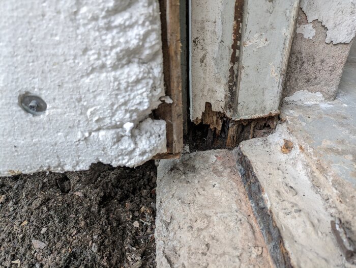 Damaged wooden door frame embedded in concrete, showing rotted wood and exposed foundation. Nearby concrete and soil are visible.