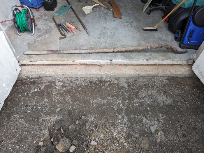 Concrete threshold area after removal, with visible tools and hose on the floor. Exposed concrete edge and rough ground below are shown.