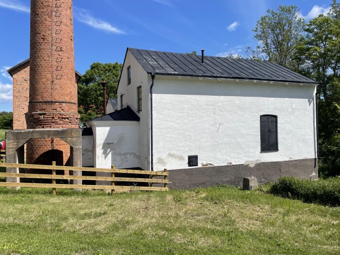Bild på en vit byggnad med svarta dörrar och ett rött tegeltorn bredvid, omgiven av gröna träd och blå himmel vid Hargs bruk.