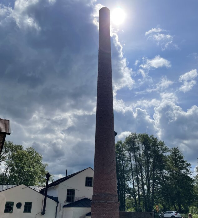 Tegelbyggnad och skorstensrör vid Hargs bruk, trädkantad bakgrund och molnig himmel med solen skymtande bakom skorstensröret.