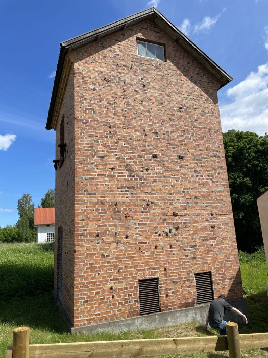 Tegelbyggnad med svart maskinsnickeri, ett fönster högt upp, två ventiler nära marken, och en person böjd nedåt. Gräs och en angränsande byggnad syns.