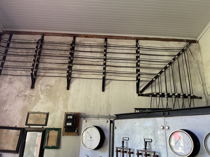 Elledningar och mätartavlor i en gammal kraftstation med synkroskop. Väggmonterade ledningar och mätare syns tillsammans med inramade dokument.