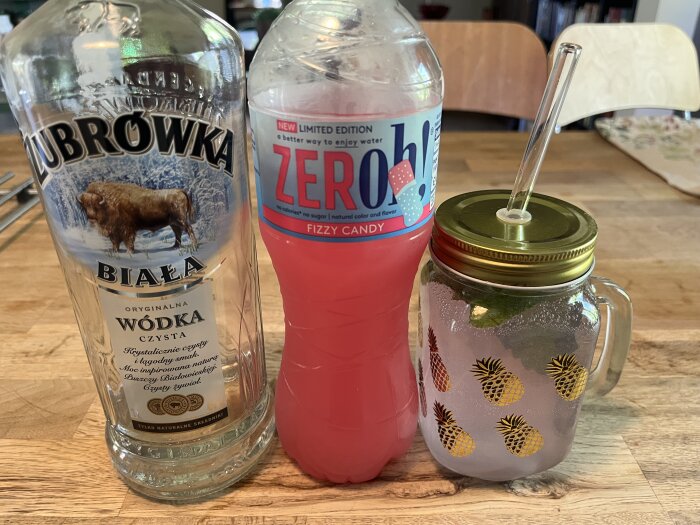 Tre föremål på ett bord: en flaska Żubrówka vodka, en flaska Zeroh! Fizzy Candy dryck och en mugg med tropiskt mönster fylld med en drink.