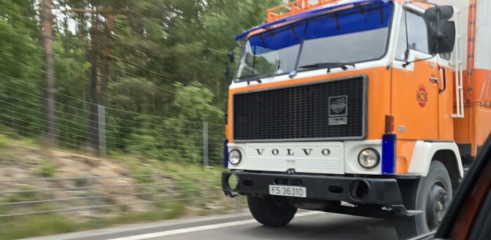 Orange och vit Volvo-lastbil på väg, E18, med omgivande skog i bakgrunden.