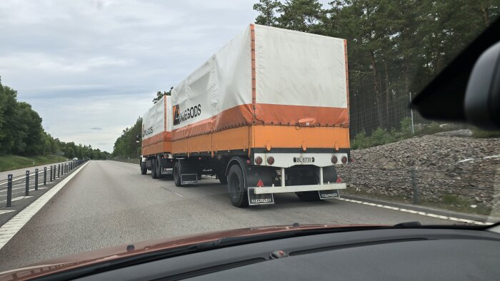 Lastbil med orange och vit presenning på släpkärran kör på E18 motorvägen, omgiven av träd och berg.