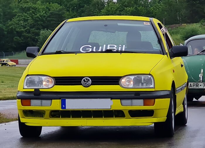 Gul Volkswagen Golf med texten "GuLBil" på framrutan parkerad på en racebana, med en grön bil i bakgrunden.