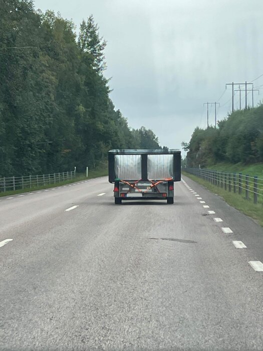 Ett stort bad fraktas på en släpkärra fastsatt med spännband, som förflyttar sig längs en landsväg med grönskande omgivning.