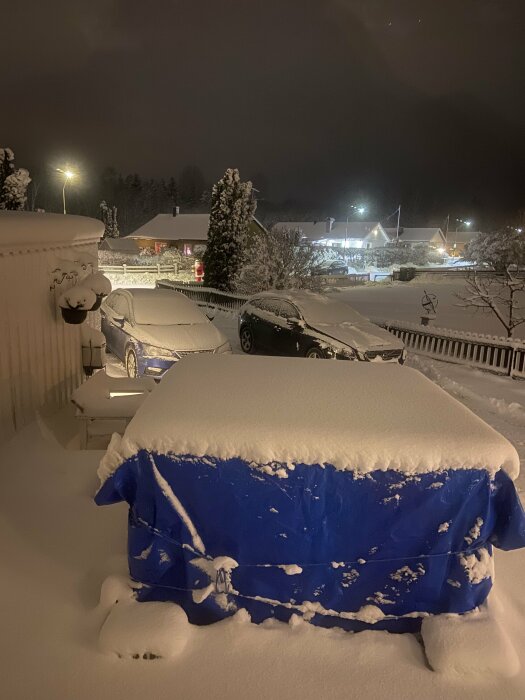 Spabad täckt med blå presenning och snö, placerat på järnrör vid sidan av ett snötäckt hus och bilar i ett vinterlandskap.