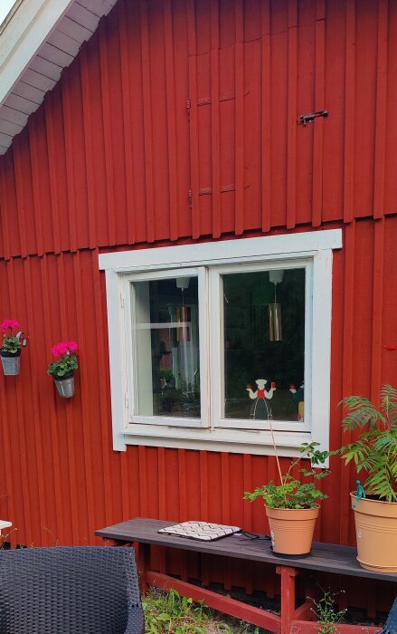 Röd husfasad med ett vitt fönster, två hängande blomkrukor, en svart korgstol, en bänk med planteringskrukor och en sittdyna.