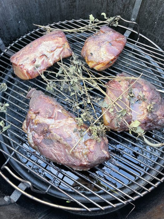 Köttstycken som röks på en grill med örter. Fyra olika bitar ligger på gallret med pinnar och kvistar ovanpå.