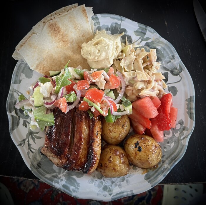 En tallrik med grillat sidfläsk, hummus, fetaostsallad, coleslaw, grillade hela potatisar, jordgubbar, melonbitar och libabröd.