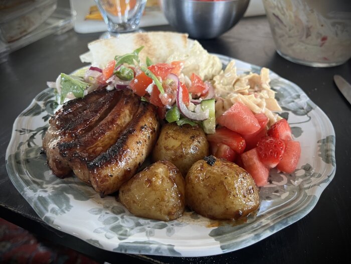 Grillat sidfläsk med stekta potatis, fetaostsallad, coleslaw, jordgubbar, melon och bröd på en tallrik.