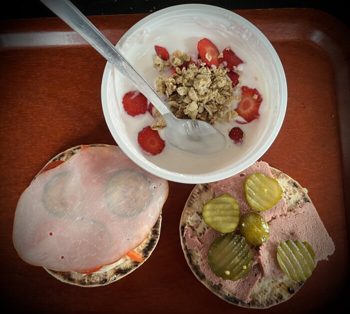 Yoghurtkvarg med färska jordgubbar och müsli, bröd med skinka och bröd med leverpastej och skivad gurka på en bricka.