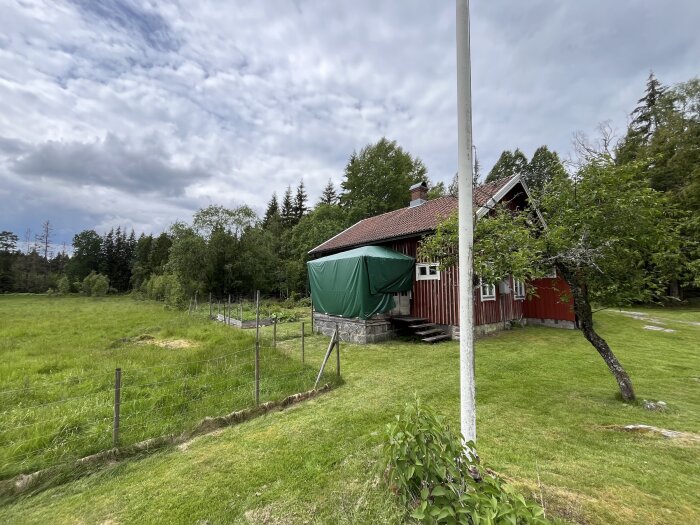 Rött timmerhus på landet med veranda i behov av renovering. Omgivet av gräsmatta och träd med skog i bakgrunden under molnig himmel.