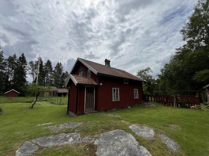 Rött timmerhus med tegeltak och veranda på landsbygden, omgiven av träd och grönska. Träfönster och dörr syns, tillsammans med en stenig gräsmatta.