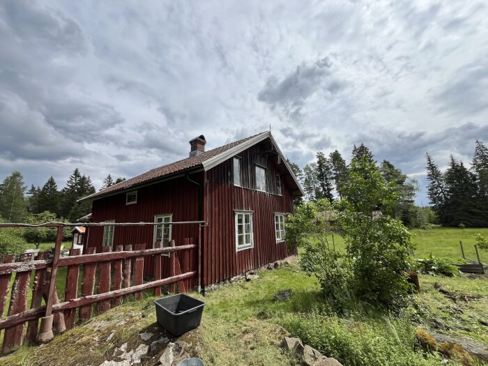 Rött timmerhus med brunt tak och vitmålade fönster. Omgivet av grönska och ett rött staket. Mörka, molniga himlen i bakgrunden.