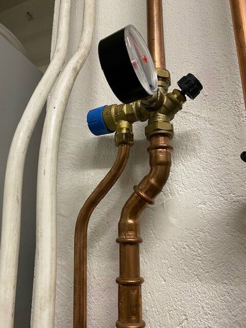 Manometer och kallvattenrör med säkerhetsventil som visar trycket 5,5 bar, i ett system med kopparledningar mot en vit vägg.