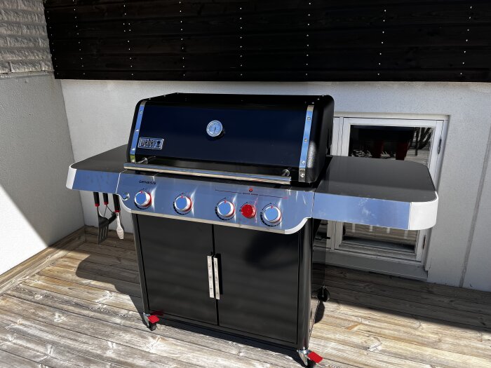 Stor grillmaskin med svart och silverfärgad design, märkt "Weber Genesis", placerad på en träbalkong. Grillsredskap hänger på vänster sida.